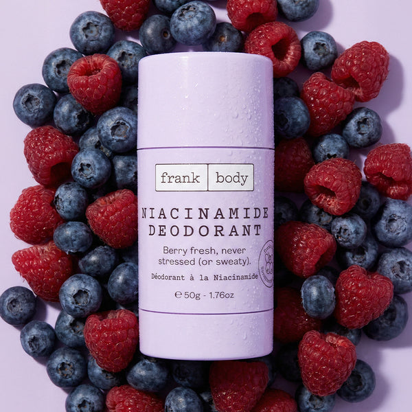Deodorant stick with 'frank body' branding surrounded by raspberries and blueberries on a light purple background
