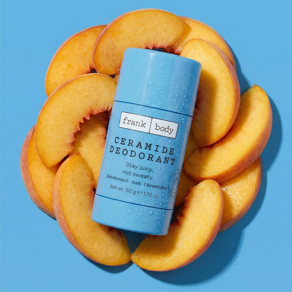 A light blue Frank Body Ceramide Deodorant stick is centered on a bright blue background, surrounded by sliced peach halves. The product label reads 'frank body CERAMIDE DEODORANT' and 'Stay juicy, not swee.