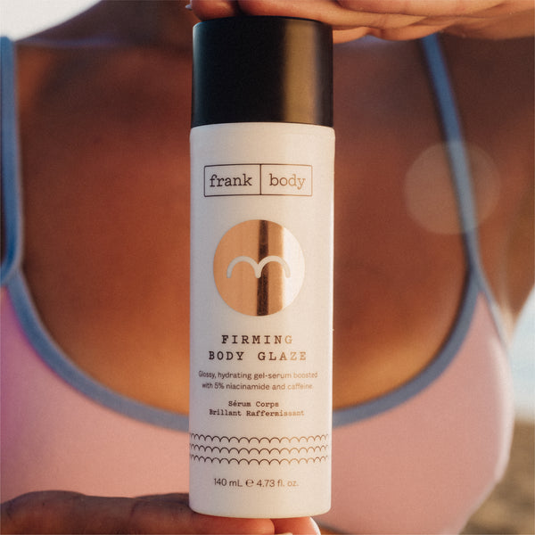 A close-up of a bottle of frank body Firming Body Glaze held against a bare back, with the label and 140 mL size legible.