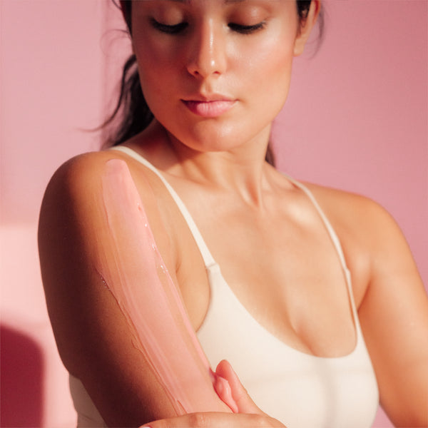 A woman in a white top with frank body Firming Body Glaze spread in a glossy pink layer down her upper arm on a pink background.