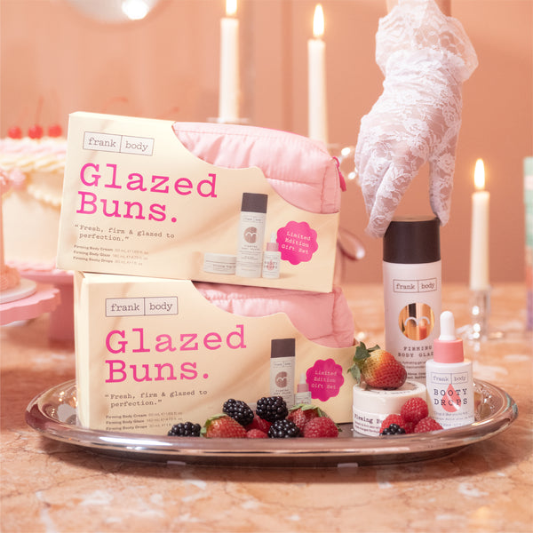 Frank Body Glazed Buns skincare set arranged on a silver tray with strawberries and blackberries, featuring two cream-colored boxes with pink lettering, a pink pouch, a white glove, a body oil bottle, and small skincare containers on a warm, candlelit background.
