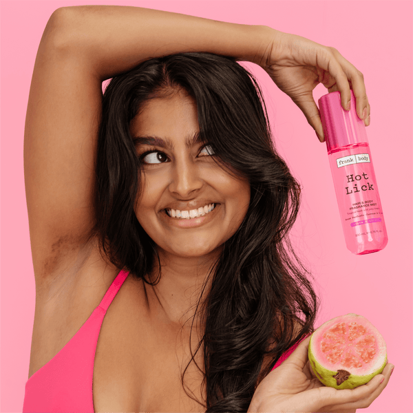 A smiling woman in a hot pink bra holding a bottle of frank body Hot Lick Hair & Body Mist and half a guava.