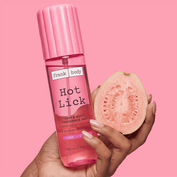 A hand holding a bottle of frank body Hot Lick Hair & Body Mist beside half a guava on a pink background.