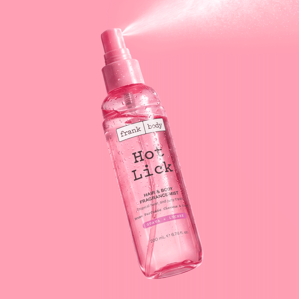 A bottle of frank body Hot Lick Hair & Body Mist spraying against a pink background.