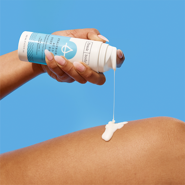 A hand dispensing frank body Hyaluronic Acid Body Serum from the bottle onto a leg against a blue background.