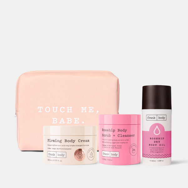 A Frank Body gift set featuring a light pink zippered pouch with white text, a beige jar of Firming Body Cream, a pink jar of Rosehip Body Scrub + Cleanser, and a white bottle of Robespay Dry Body Oil with a dark brown cap, arranged on a light gray background.