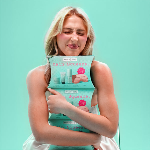 A blonde woman in a white dress hugs a stack of Frank Body Main Squeeze hand towels against a solid mint green background.
