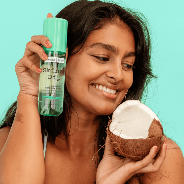 A smiling woman holding a bottle of frank body Skinny Dip Hair & Body Mist and half a coconut.