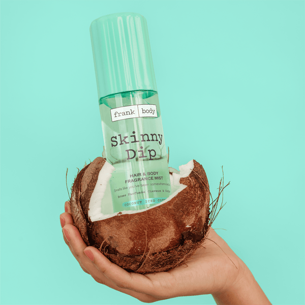 A bottle of frank body Skinny Dip Hair & Body Mist resting in half a coconut held in a hand.