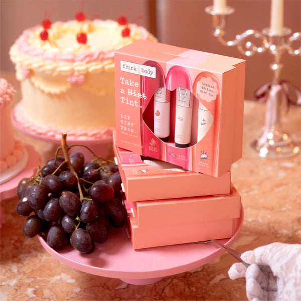 A pink boxed hair tint set labeled 'frankie body' sits on a pink cake stand in a styled indoor scene. The set includes three cosmetic tubes visible through a window in the box, with additional matching boxes stacked below.