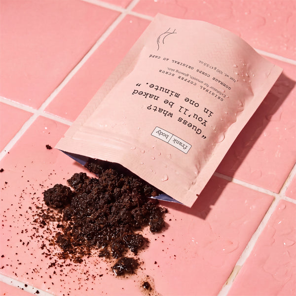 A pink pouch labeled lies open on pink tiled flooring, with dark coffee grounds spilled out. Water droplets are visible on the pouch and surrounding surface.
