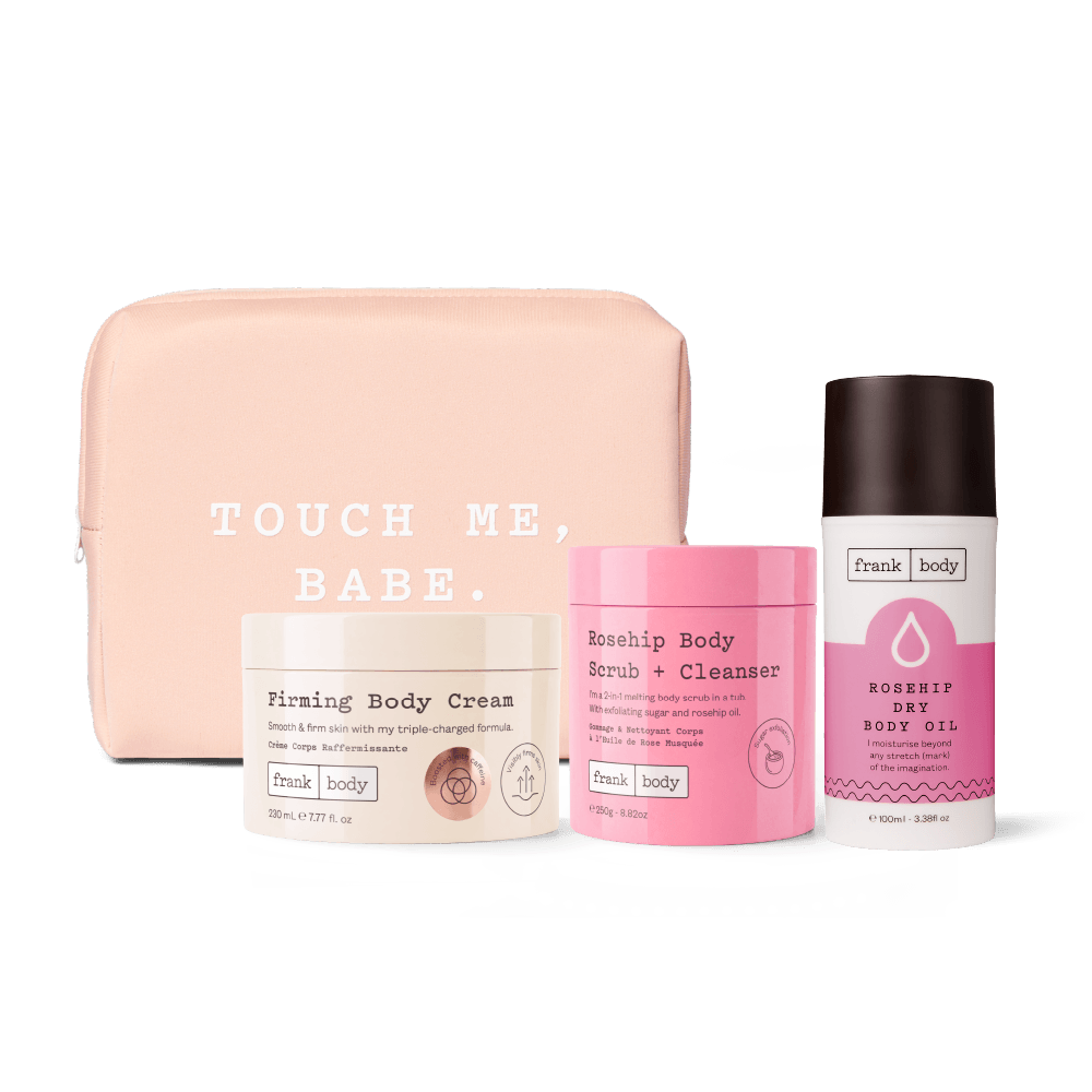 A set of Frank Body self-care items arranged in front of a blush pink zip pouch. The set includes a jar of Firming Body Cream, a tub of Rosehip Body Scrub + Cleanser, and a deodorant-style bottle of Rosehip Dry Body Oil.