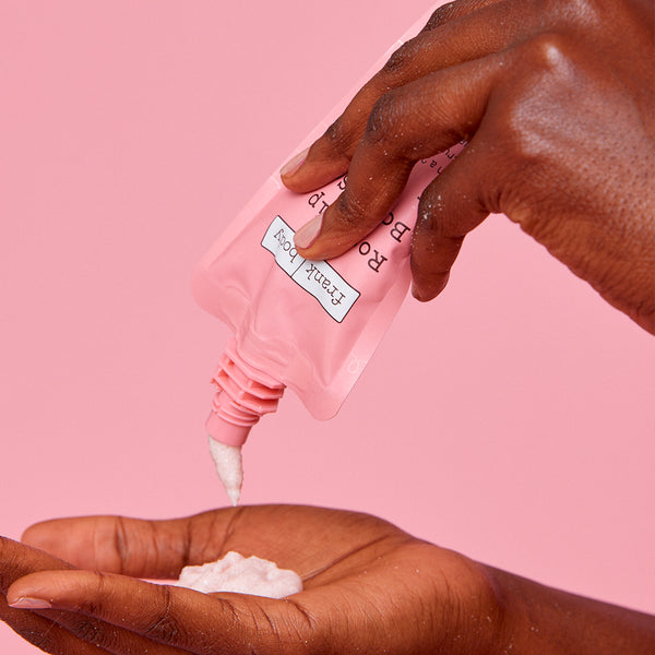 A close-up of a hand squeezing a pink squeeze pouch labeled 'Rosehip Body Scrub' to dispense white cream into another hand against a solid pink background.