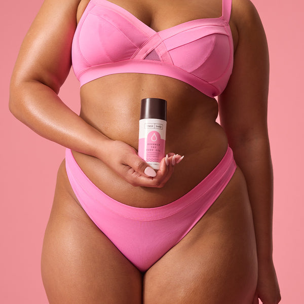 A smiling woman in a pink bra and underwear holding a bottle of frank body Rosehip Dry Body Oil.