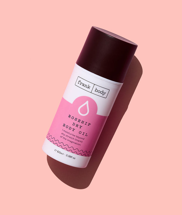 A bottle of frank body Rosehip Dry Body Oil with a dark cap on a pale pink background.